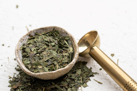 A bowl filled with Guradji tea leaves accompanied by a shiny metal spoon resting beside it.