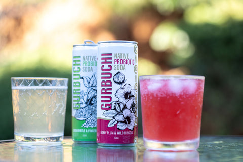 Two cans of Gurbuchi Native Probiotic Soda beside two glassed filled with fresh Gurbuchi, showcasing vibrant colours and refreshing appeal.